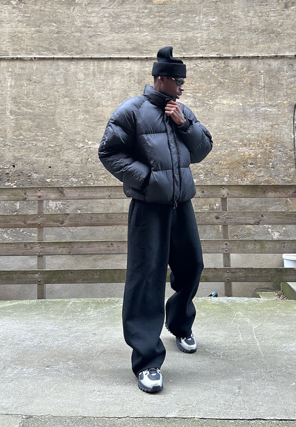 Black puffer jacket with a high collar, paired with loose black trousers. The outfit is completed with metallic silver sneakers and a black knit beanie.