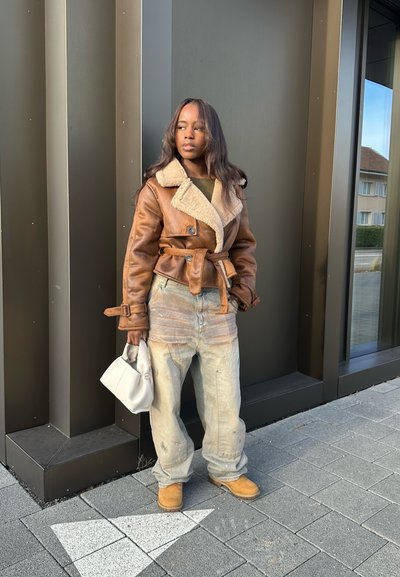 Chaqueta de cuero marrón con cuello de borrego, ceñida en la cintura. Lleva pantalones holgados de color claro y botas marrones. Sostiene un bolso blanco.