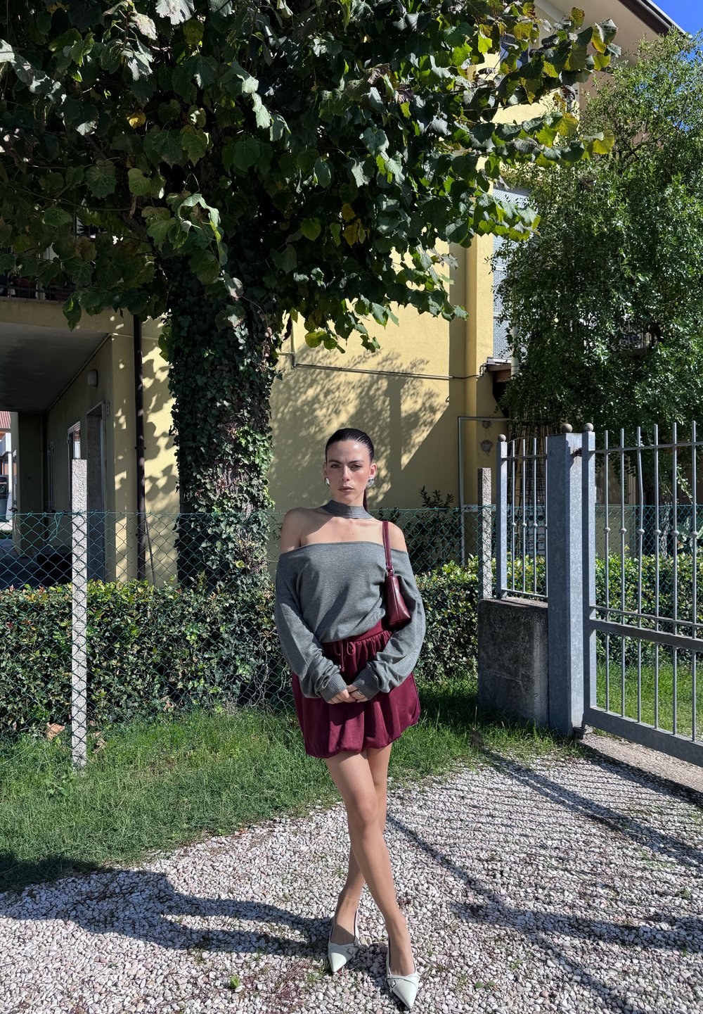 Avtagande axel grå tröja, vinröd kjol, bleka gröna klackar och matchande väska; står i ett grönområde med träd i bakgrunden.