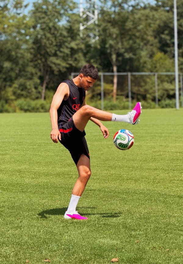 Jeune homme en tenue de sport noire jonglant avec un ballon de football sur un terrain vert avec des arbres et un but en arrière-plan par une journée ensoleillée.