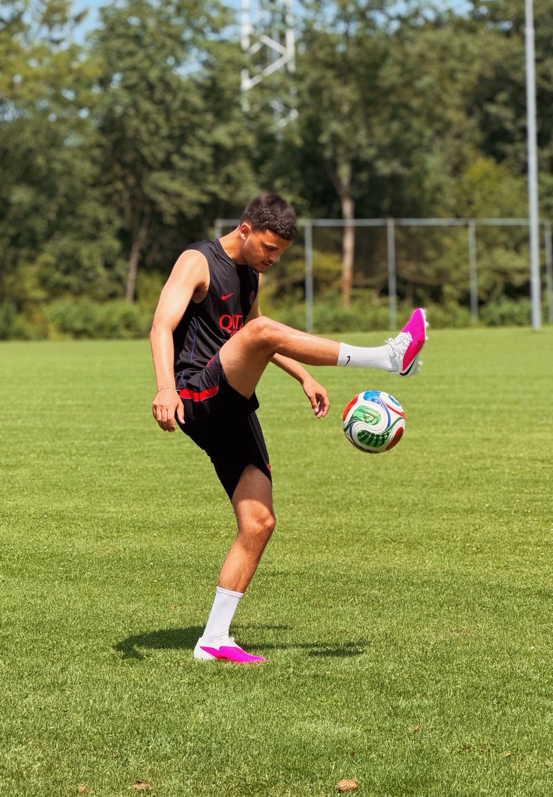 Jeune homme en tenue de sport noire jonglant avec un ballon de football sur un terrain vert avec des arbres et un but en arrière-plan par une journée ensoleillée.