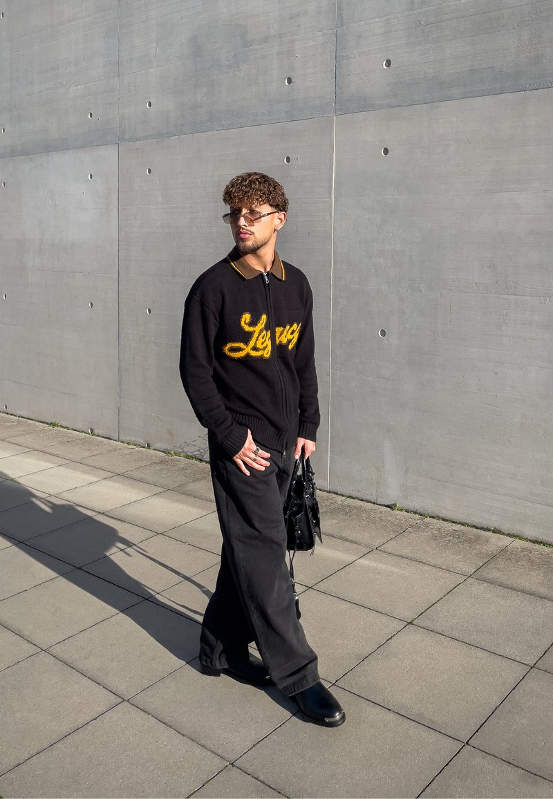 Sweater preto com fecho éclair e logotipo amarelo "Levi's", colarinho castanho, calças largas pretas, sapatos de couro pretos e uma mala preta. Fundo texturizado.