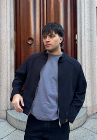 Jeune homme aux cheveux courts et foncés portant une veste noire sur un t-shirt gris, debout devant une porte en bois encadrée de colonnes en pierre.