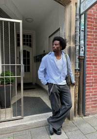 Ανοιχτό μπλε ριγέ πουκάμισο, λευκή μπλούζα από κάτω, γκρι φαρδιά τζιν παντελόνια με λεπτομέρεια αλυσίδας, και μαύρα παπουτσοειδή, στέκεται δίπλα σε μια πύλη.