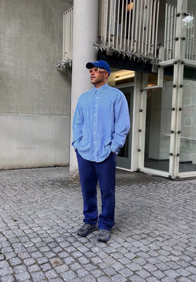 Camisa de botones a rayas azul y blanca, pantalones azules, zapatillas grises, gorra azul y gafas de color claro, de pie sobre un pavimento de adoquines.