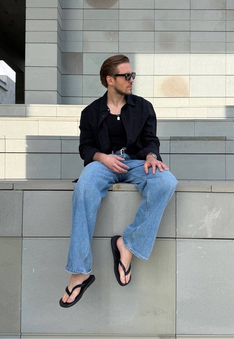 Homme portant des lunettes de soleil, une chemise noire et un jean bleu assis sur des marches en béton avec un mur carrelé gris en arrière-plan.