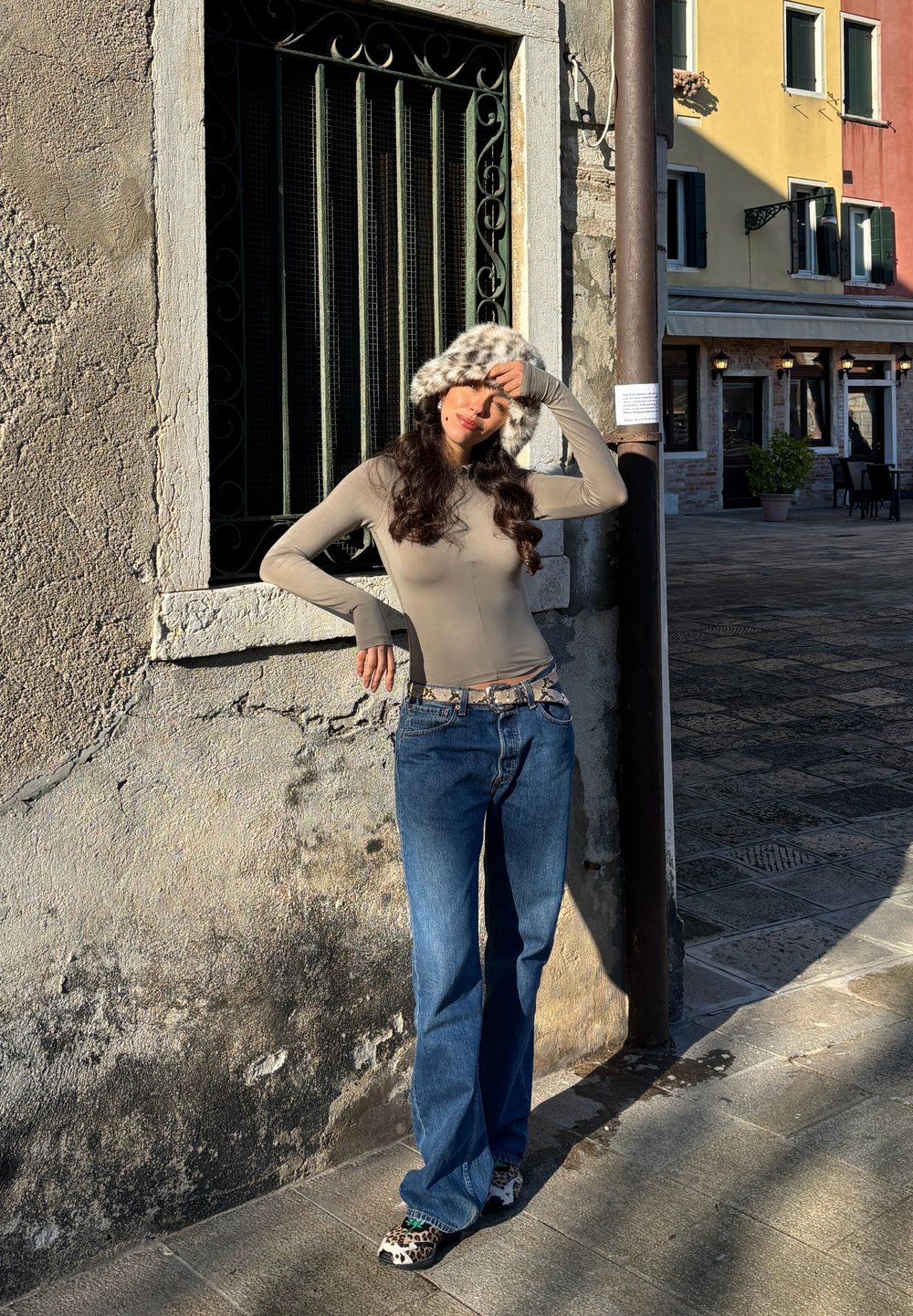 Beige enganliegendes Langarmshirt, blaue Schlagjeans und ein Kunstpelzhut mit gesprenkeltem Muster. Stehend vor einer strukturierten Steinmauer.