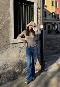 Beige enganliegendes Langarmshirt, blaue Schlagjeans und ein Kunstpelzhut mit gesprenkeltem Muster. Stehend vor einer strukturierten Steinmauer.