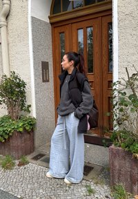 Pilka per didelis megztinis, juoda pūkinė liemenė ir šviesiai pilkos plačios kelnės. Ruda petnešų rankinė, stovinti prie medinių durų su stiklinių panelių.