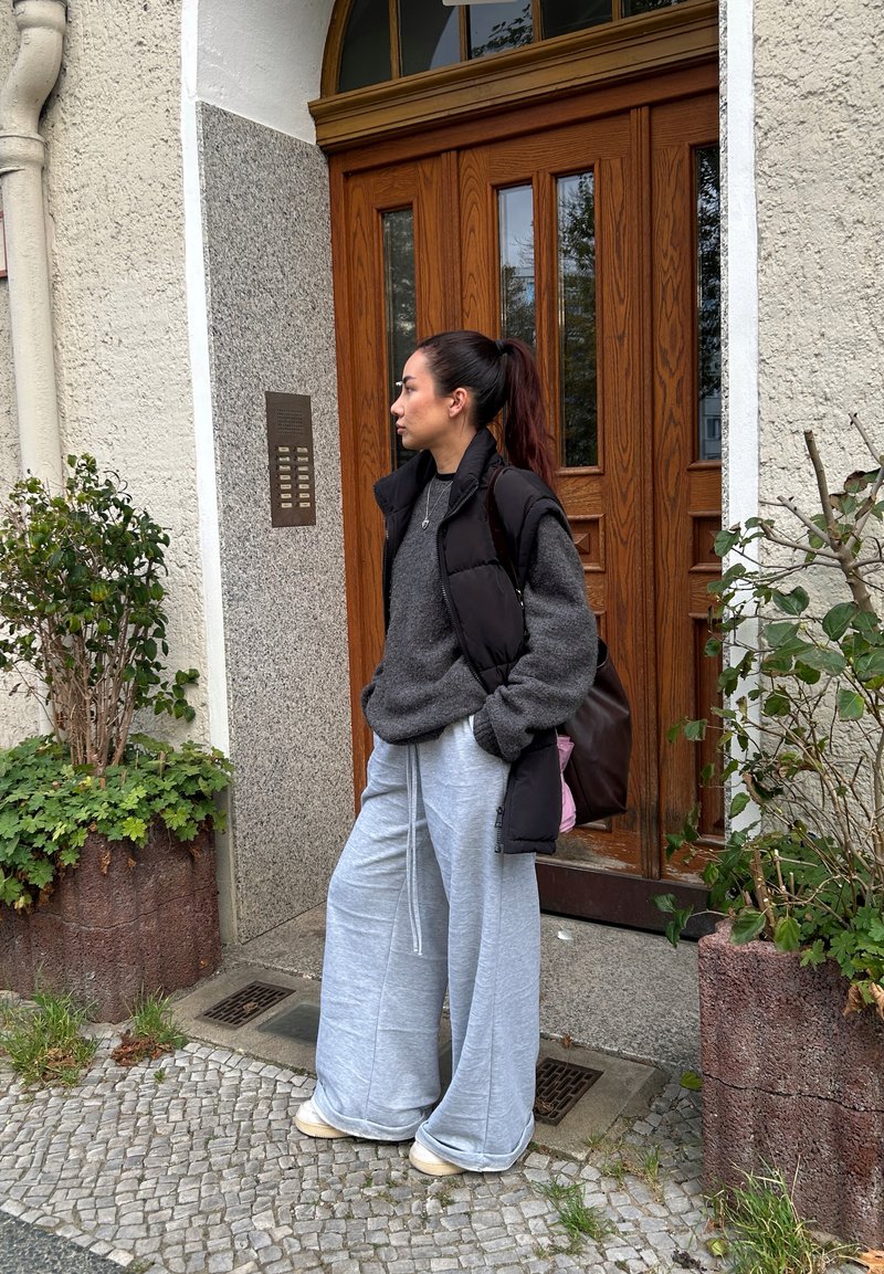 Grå oversized tröja, svart puffer väst och ljusgrå vida sweatpants. Brun axelväska, stående vid en trädörr med glaspartier.