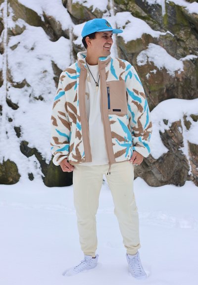 Hombre con gorra azul, chaqueta de forro polar estampada, pantalones blancos y zapatillas de deporte, de pie sobre la nieve frente a un terreno rocoso cubierto de nieve.