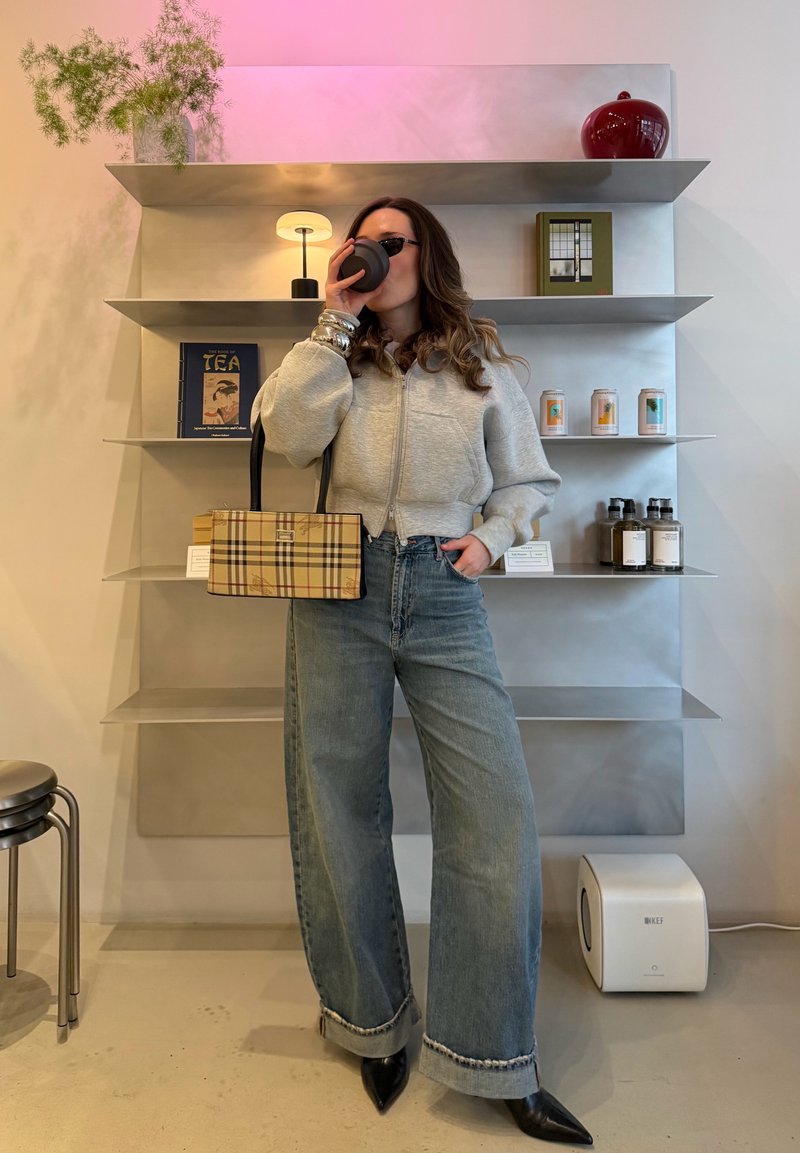 Femme portant des lunettes de soleil et un sweat-shirt gris à fermeture éclair, buvant dans une tasse, tenant un sac à main à carreaux, dans une pièce moderne avec des étagères.