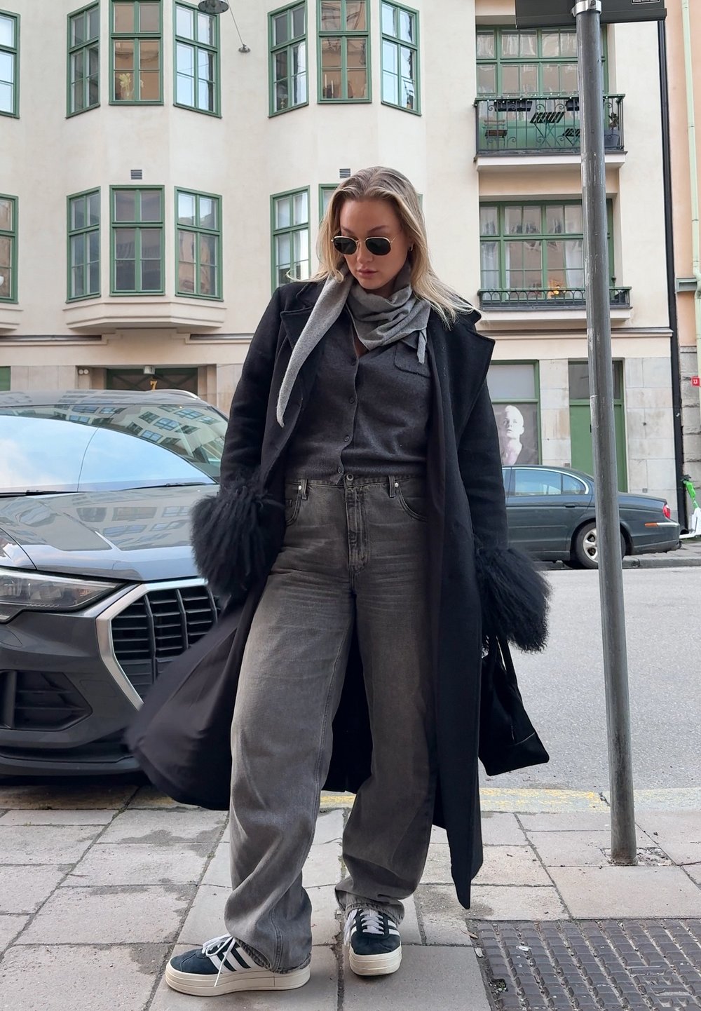 Femme portant un manteau à revers en fourrure noire, une écharpe grise, un cardigan gris foncé, un jean gris large et des baskets, debout sur un trottoir urbain près de voitures garées.