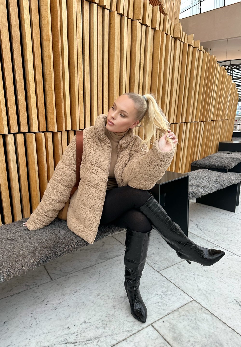 Beige, fluffig teddyjacka med hög krage, matchad med en brun polotröja och svarta leggings. Glansiga svarta knähöga stövlar. Träbakgrund.