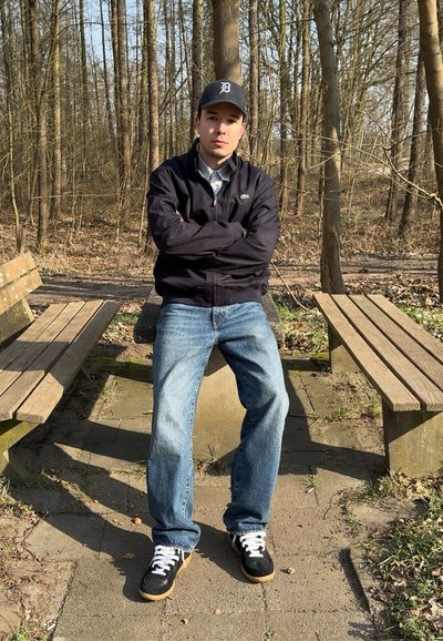 Persona sentada en un banco de piedra, llevando una chaqueta negra de cremallera, una camisa clara, jeans azules y zapatillas negras con detalles blancos en un área boscosa.