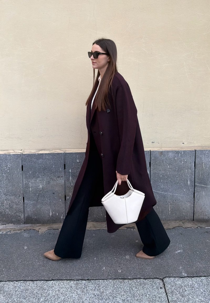 Femme marchant sur le trottoir portant un long manteau sombre, un pantalon large noir, des lunettes de soleil, des chaussures camel, et portant un sac à main blanc devant un mur beige.