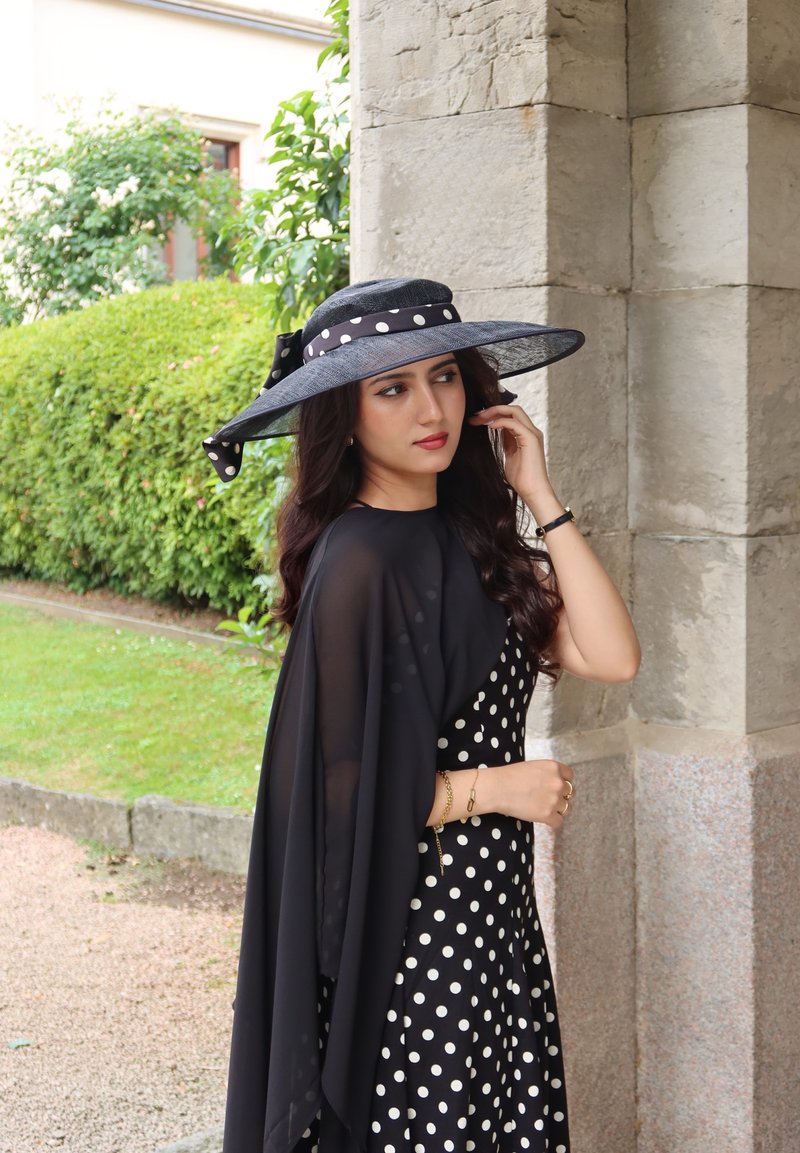 Woman in black dress with white polka dots and wide-brimmed hat stands near stone column, touching her hair outdoors by green bushes.