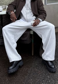 Pantalon de jogging gris à coupe ample, associé à des chaussures plateformes noires épaisses ; assis à une table, tenant une petite tasse blanche.