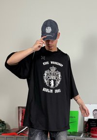Must värviline oversize särk valge printimisega, kus on kujutatud krooni ja tekst "BERLIIN 2001". Hall müts embleemiga ja lõdva istuvusega. Tekstuuriga tumedad mustrilised püksid.
