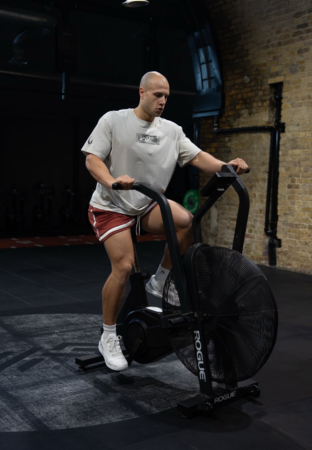 Svart vifte-sykkel med en stor sirkulær vifte, robust ramme og justerbare styrehåndtak. Bruker iført lys skjorte, røde shorts og hvite joggesko.