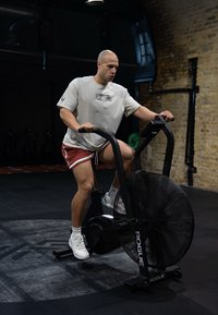 Svart vifte-sykkel med en stor sirkulær vifte, robust ramme og justerbare styrehåndtak. Bruker iført lys skjorte, røde shorts og hvite joggesko.