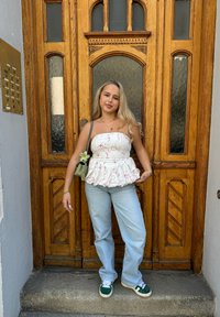 Weißes Peplum-Oberteil mit roten Wirbeln, hellblaue Jeans und grüne Sneakers. Steht neben einer Holztür mit Glasfenstern. Lässiger Stil.