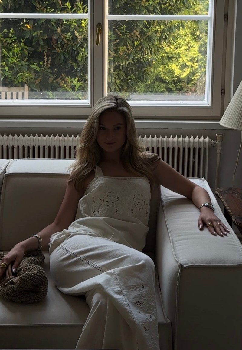 Vrouw in witte jurk ligt ontspannen op een beige bank bij een raam met groen buiten, met een gevlochten tas op haar schoot.