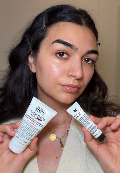 Tubes del Limpiador Facial Ultra de Kiehl's y del Bálsamo Labial sostenidos por una persona, con un envase blanco y texto negro y acentos rojos, sobre un fondo claro.