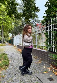 Ljusbeige kort jacka över en rosa topp, svart ribbad kjol och svarta snörstövlar med vita detaljer. Fortfarande på trottoaren med grönska i bakgrunden.