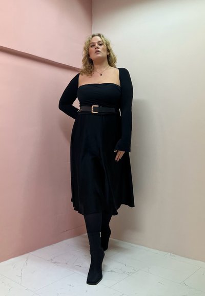 Vestido negro de hombros al descubierto con mangas largas, cintura ceñida con un cinturón y una silueta fluida. Llevado con botas negras de tacón.