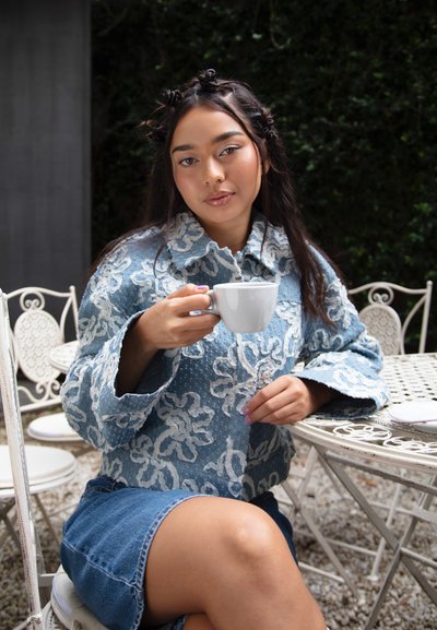 Chaqueta azul con patrón floral, llevada sobre una falda de denim azul. Sosteniendo una taza blanca, sentada en una mesa de metal blanco.