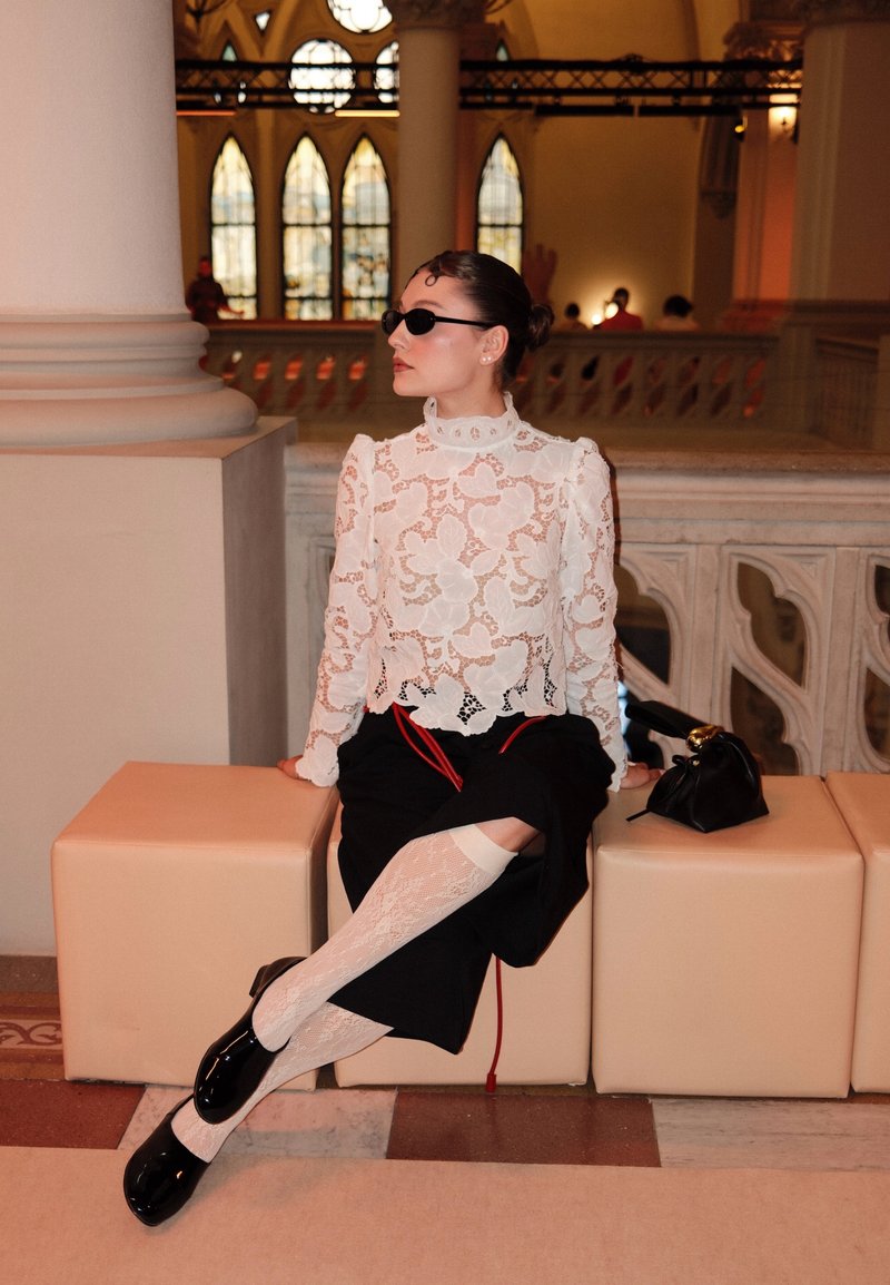 Femme portant un haut en dentelle blanc, une jupe noire, des bas blancs et des chaussures noires, assise sur des bancs beige à l'intérieur d'un bâtiment avec des vitraux.
