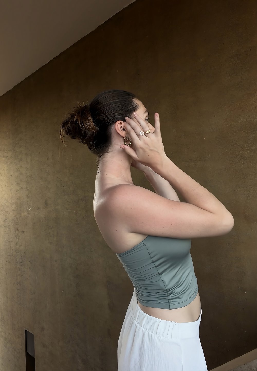 Femme aux cheveux attachés en chignon, portant un haut bleu sans manches et une jupe blanche, ajustant ses boucles d'oreilles devant un mur marron uni.