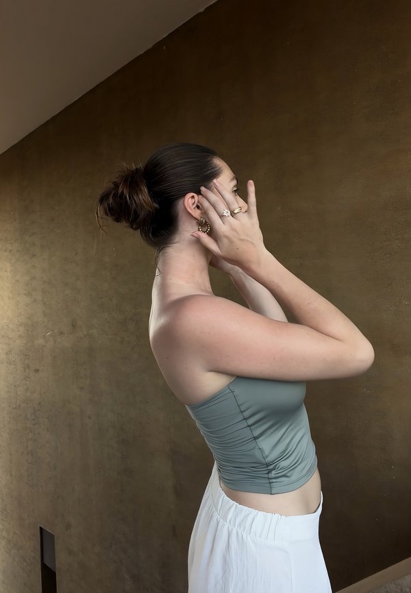 Ženska z lasmi v figi, oblečena v modro brezrokavnik in belo krilo, si proti enostavni rjavi steni prilagaja uhane.