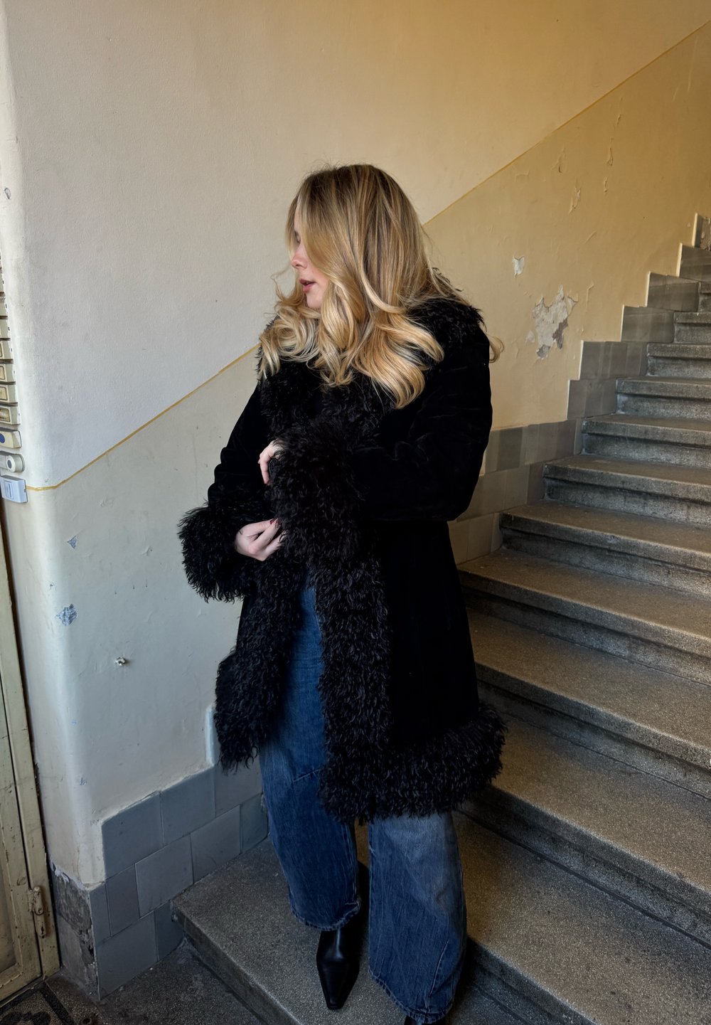 Manteau noir doublé de fourrure avec des poignets texturés, porté sur un jean large bleu et des bottines noires, debout sur un escalier avec un mur texturé.