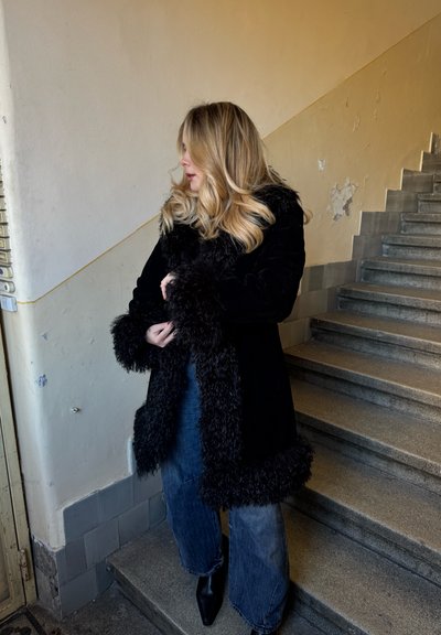 Abrigo negro forrado de piel con puños texturizados, llevado sobre unos vaqueros azules de pierna ancha y botines negros, de pie en una escalera con una pared texturizada.