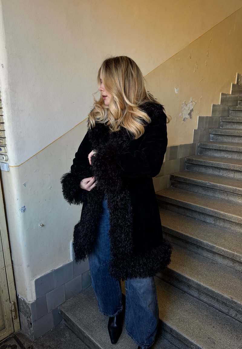 Svart pelsforet frakk med teksturerte mansjetter, båret over blå vidbente jeans og svarte ankelstøvler, stående på en trapp med en teksturert vegg.