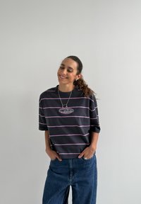 T-shirt oversize rayé noir et violet avec manches courtes et col rond, arborant un logo graphique central. Porté avec un jean bleu ample.