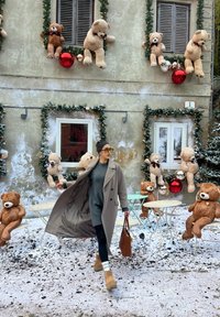 Femme en manteau d'hiver et bottes marchant devant un café en plein air enneigé décoré d'ours en peluche, de guirlandes et de grandes décorations rouges et argentées.