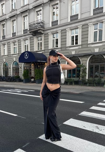 Top corto negro con detalles blancos, falda negra con abertura alta y nudo, bolso plateado, gorra negra, zapatos negros con tiras, entorno urbano.