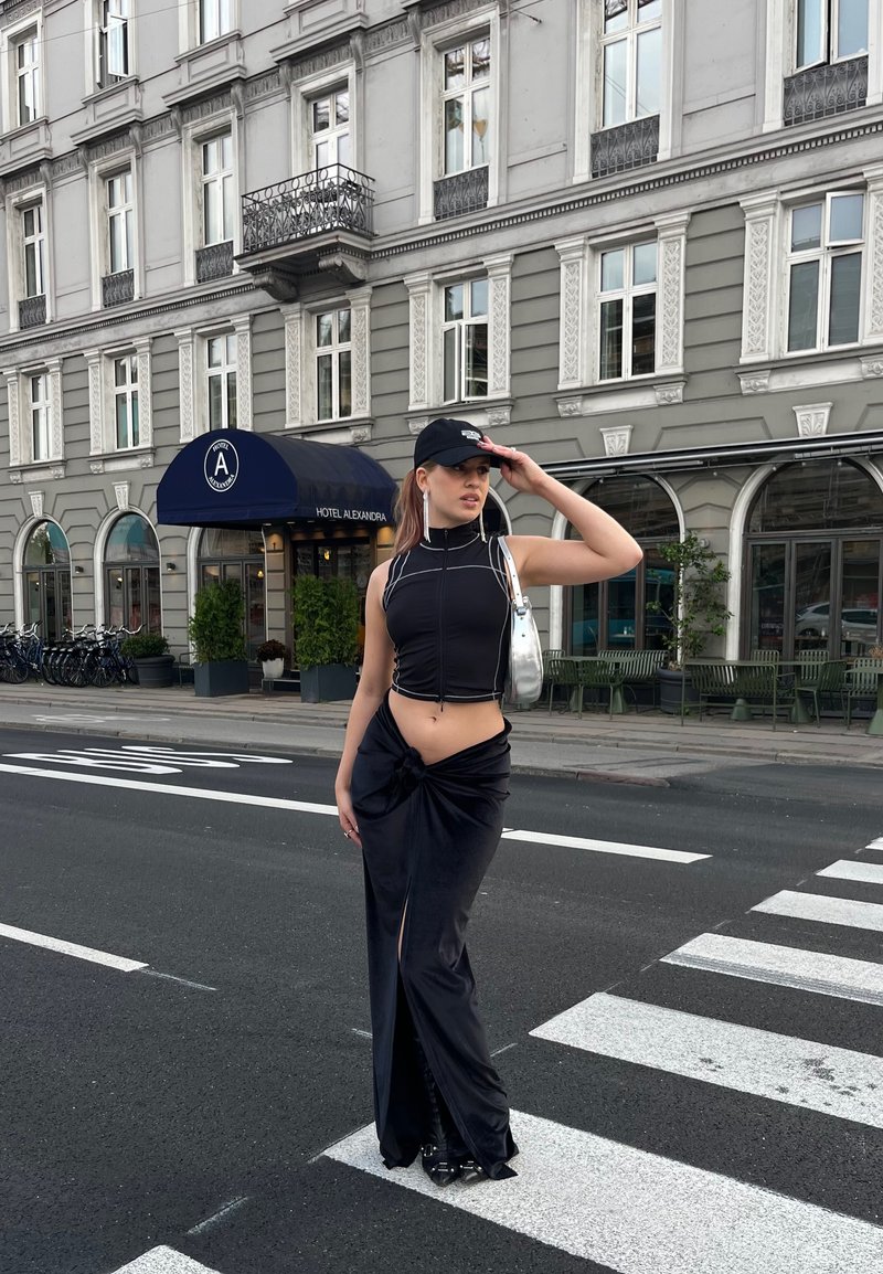 Μαύρο κοντό τοπ με λευκές λεπτομέρειες, μαύρη φούστα με ψηλή σχισμή και κόμπο, ασημένια τσάντα, μαύρο καπέλο, μαύρα σανδάλια με λουριά, αστική τοποθεσία.