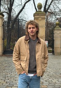 Veste en toile de coton beige avec col marron, associée à un pull foncé et un jean bleu. Les caractéristiques remarquables incluent des poches et une coupe décontractée.