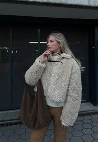 Κρεμ απόχρωσης φουσκωτό τζάκετ από σέβερ με κουκούλα, κλείσιμο με κουμπιά και ριγωτό στρίφωμα. Συνδυασμένο με καφέ παντελόνια και μια καφέ σουέντ τσάντα.