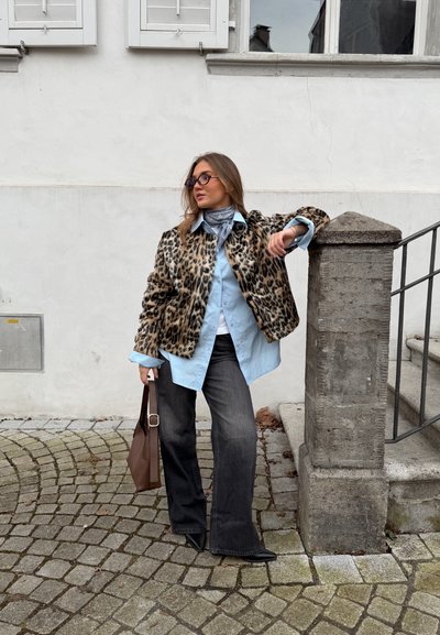 Mujer con chaqueta de estampado de leopardo, camisa azul claro, jeans oscuros y gafas de sol, apoyada en un pilar de piedra junto a unas escaleras al aire libre, sosteniendo una bolsa marrón.