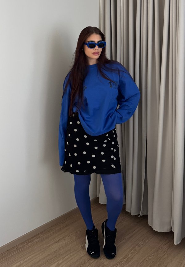 Woman wearing blue sunglasses, blue long-sleeve top, black skirt with white polka dots, blue tights, and black sneakers standing indoors.