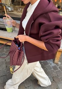 Bordeaux hoodie met grote mouwen; crèmekleurige top; gevarieerde geweven bordeaux tas; houdt een heldere drank met een rietje vast.