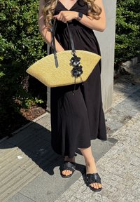 Femme en robe noire tenant un grand sac en paille avec des décorations de fleurs noires, portant des sandales noires avec les ongles des orteils peints en jaune.