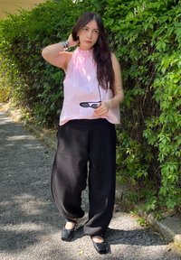 Top sem mangas rosa claro com decote franzido, combinado com calças largas pretas e sapatos pretos. Modelo segura óculos de sol escuros. Fundo verde.