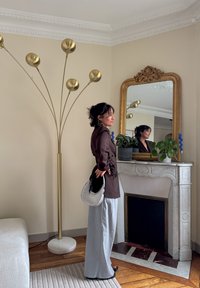 Haut marron à manches longues à enfiler, pantalon gris à jambes larges, et sac à main rond en argent. Lampe sur pied dorée, grand miroir orné, et plantes vertes en pots.