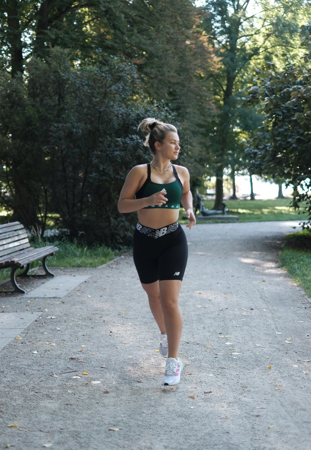 Mladá žena se běhá na štěrkové cestě v parku, má na sobě zelenou sportovní podprsenku, černé šortky a bílé tenisky.